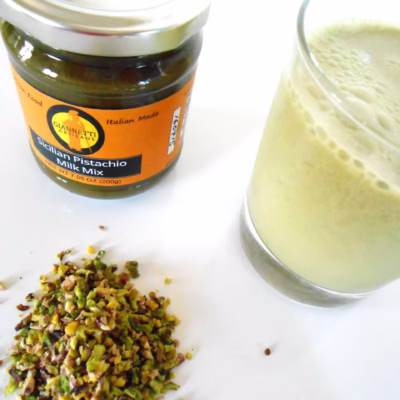 Top view of 7 oz Giannetti Artisans Pistachio Paste jar and a glass of the pistachio milk with pistachio grains on the side on a white background